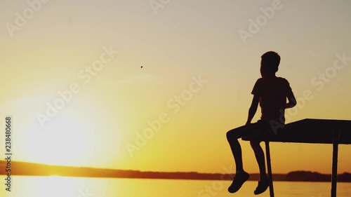 A little child is sitting on the bridge above the lake and throwing stones into the water at sunset in the summer on a warm evening.