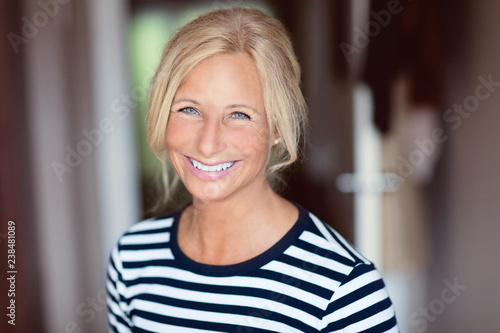 Middle age woman smiling. She is at home. In a bedroom wearing a striped sweater