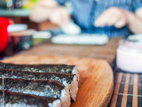 Making Sushi Rolls: Rolls Sealing On the Wooden Board before Slicing. Blurred Woman Hands Putting Rice on a Nori Sheet and Kitchenware in Background. photo