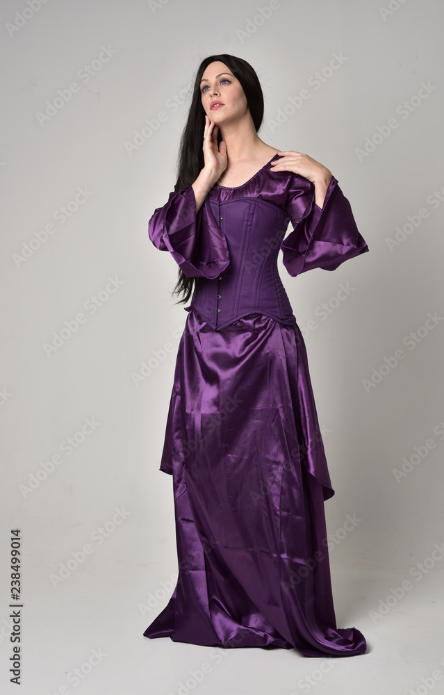 full length portrait of beautiful girl with long black hair,   wearing purple fantasy medieval gown. standing pose on grey studio background.
