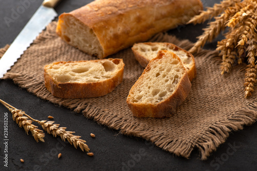 Italian traditional ciabatta bread with sliced ​​pieces and ears of wheat lie on a jute napkin on a table.  Next is a knife