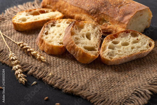 Italian traditional ciabatta bread with sliced ​​pieces and ears of wheat lie on a jute napkin on a table.