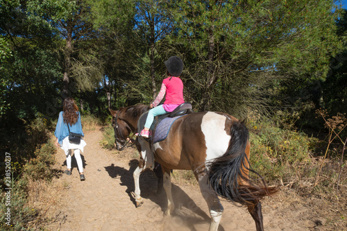 view from back of little four years old girl riding a horse next to her mother walking in a forest