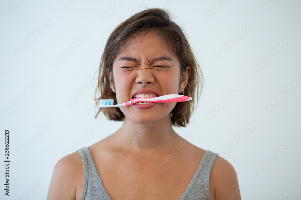 Portrait of mischievous young woman holding toothbrush in teeth. Frowning Asian girl during morning routine. Dental care concept Portrait of mischievous young woman holding toothbrush in teeth. Frowning Asian girl during morning routine. Dental care concept