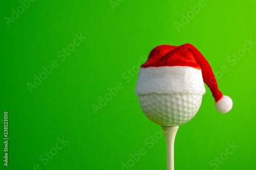 Golf ball in a red Santa Claus hat mounted on a tee. Sports concept on the theme of Christmas and New Year. Green background. Copy space.