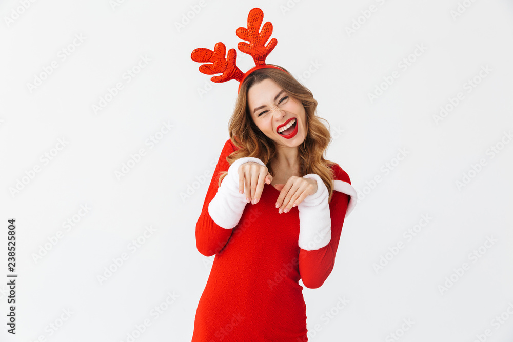 Portrait of flirting woman 20s wearing Santa Claus red costume and deer ears smiling and standing, isolated over white background Portrait of flirting woman 20s wearing Santa Claus red costume and deer ears smiling and standing, isolated over white background