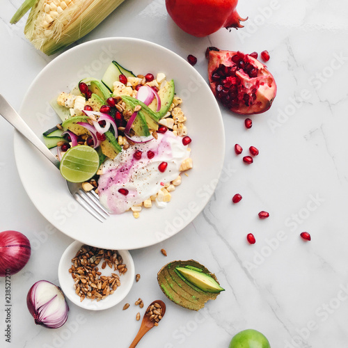 Plate of colorful healthy salad with avocado, sweet corn, pomegranate, cucumber, red onion, lime, greek yogurt and sunflower seeds on a marble background, top view