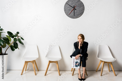 bored young businesswoman holding newspaper and sitting with hand on chin in queue