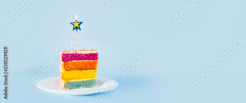Slice of Rainbow cake with birning candle in the shape of star on white round plate isolated on blue background. Happy bithday, party concept. Wide banner. Selective focus. Copy space.
