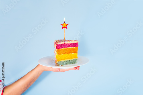Female hand holding white plate with slice of Rainbow cake with birning candle in the shape of star isolated on blue background. Happy bithday, party concept. Square card. Selective focus. Copy space.