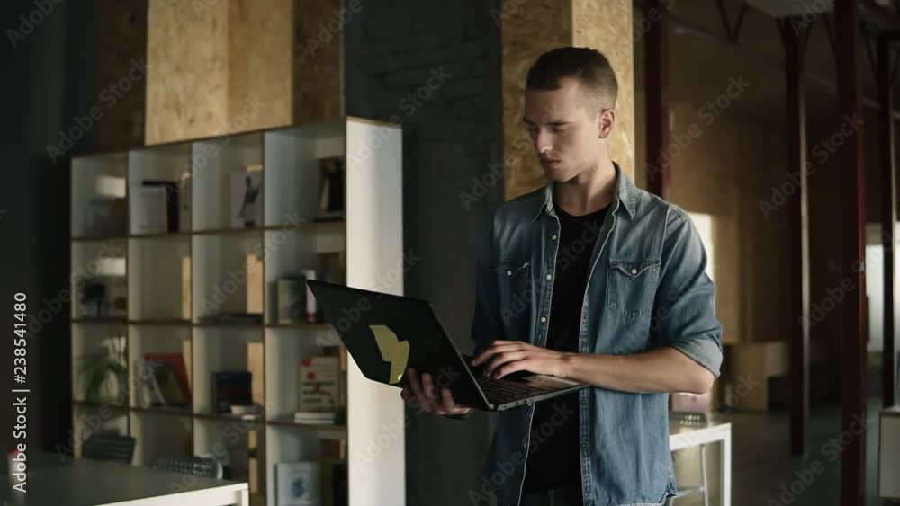 Attractive concentrated young business man is walking with a laptop in his hand and typing. Caucasian positive man working in public workplace with brick wall interior. Slow motion. Front view Attractive concentrated young business man is walking with a laptop in his hand and typing. Caucasian positive man working in public workplace with brick wall interior. Slow motion. Front view