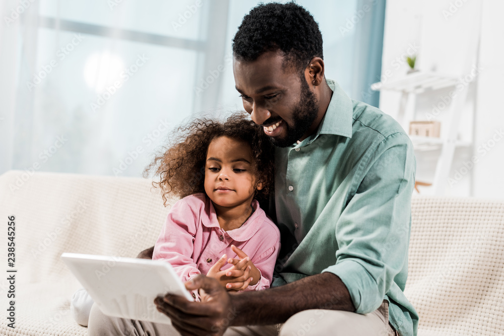 Obraz premium smiling african american father and kid using digital tablet while sitting on couch