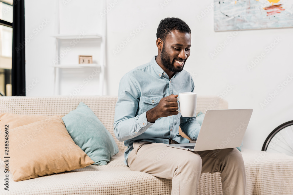 smiling african american freelancer drinking tea and using laptop in living room smiling african american freelancer drinking tea and using laptop in living room