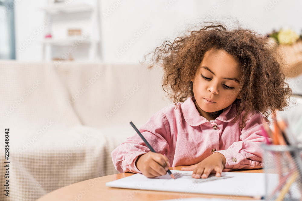 serious african american kid drawing on paper with color pencil in living room