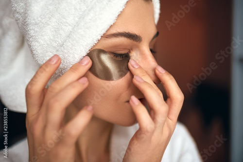 Closeup indoor portrait of beautiful woman applying eye patches on the area under the eyes from wrinkles and dark circles. Attractive female in white bathrobe making beauty treatments at home.
