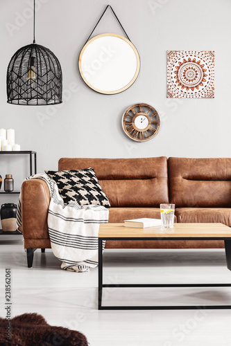 Round mirror, wooden clock and ethno graphic on the beige wall of elegant living room interior with leather couch with patterned pillow and stylish black chandelier, real photo