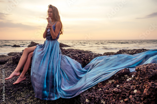 Beautiful brunette girl in blue gray chameleon dress with long train sitting on a beach at amazing sunset. woman in chic outfit near a rock on a tropical paradise island enjoying solitude and freedom
