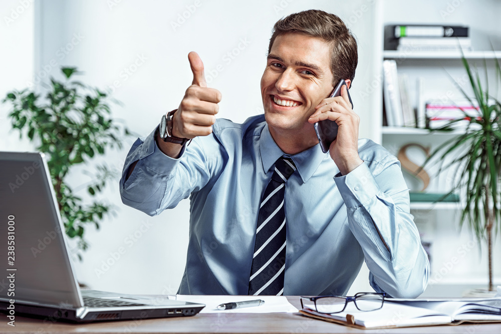 Cheerful businessman talking on the phone and showing thumb up. Photo of successful man working in the office. Business concept Cheerful businessman talking on the phone and showing thumb up. Photo of successful man working in the office. Business concept