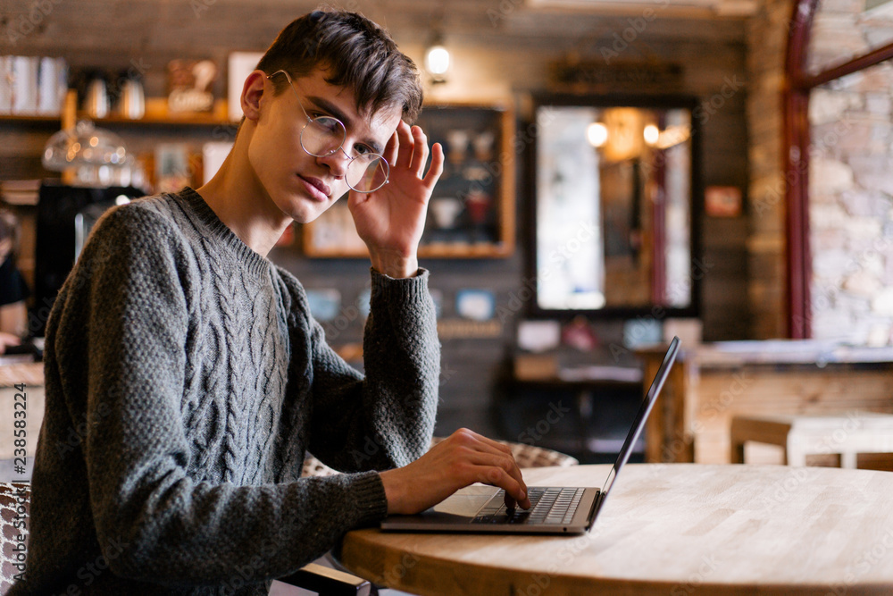 Stylish guy with glasses sitting in a cozy cafe at the laptop typing looks towards the camera Stylish guy with glasses sitting in a cozy cafe at the laptop typing looks towards the camera