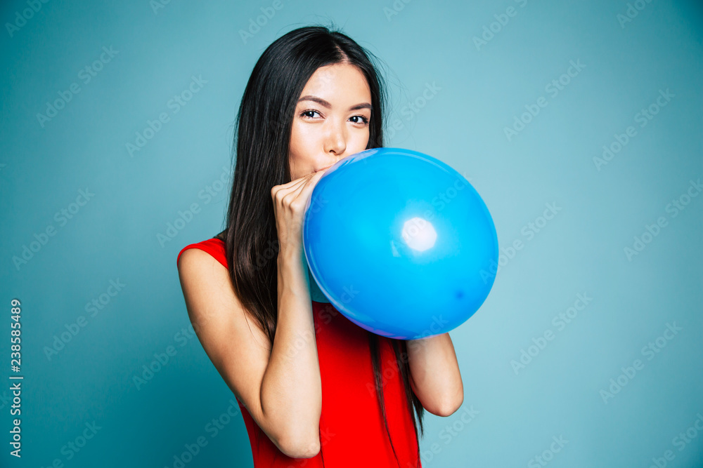 Happy emotional young asian woman in red is blowing a blue balloon in studio. Portrait of beautiful chinese girl. Shopping concept Happy emotional young asian woman in red is blowing a blue balloon in studio. Portrait of beautiful chinese girl. Shopping concept