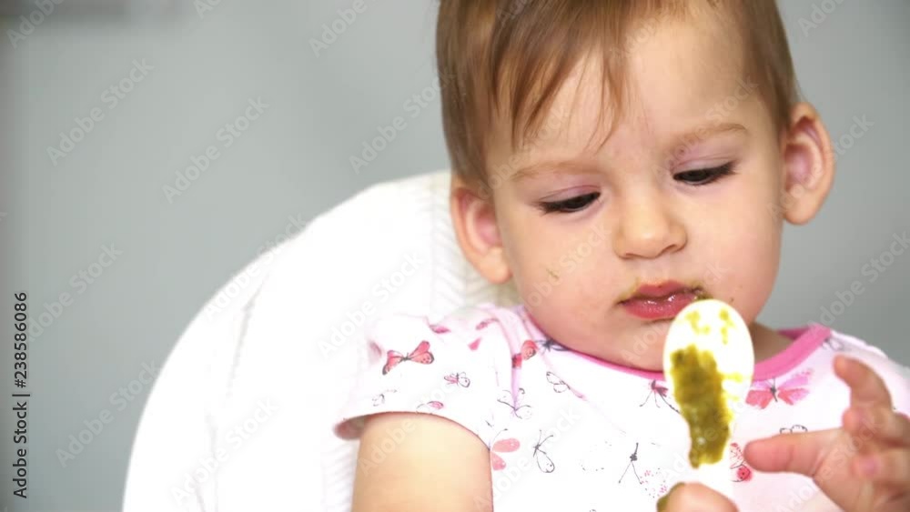 Cute baby girl holding a spoon with vegetables. A child studies spoon, vegetables fall from spoon on table Cute baby girl holding a spoon with vegetables. A child studies spoon, vegetables fall from spoon on table