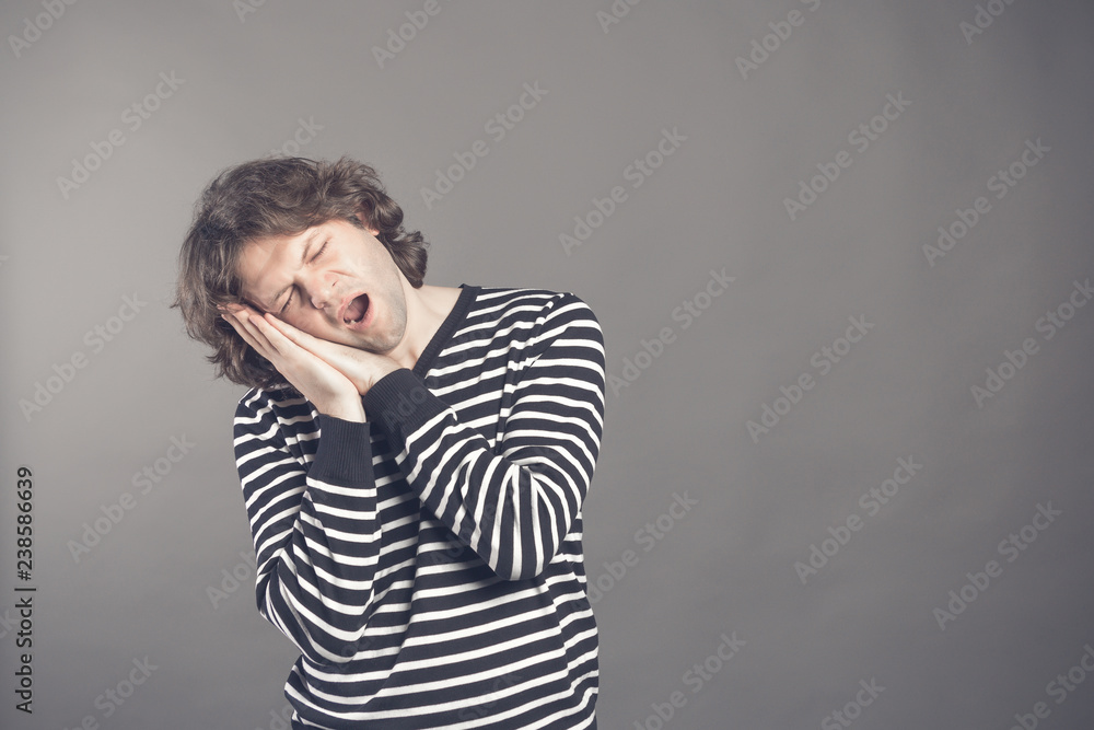 Young man in striped black and white sweater wants to sleep. Put his head in his hands and closed eyes while yawning. Standing on the grey background. Hands under his cheek. Insomnia concept Young man in striped black and white sweater wants to sleep. Put his head in his hands and closed eyes while yawning. Standing on the grey background. Hands under his cheek. Insomnia concept