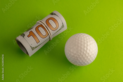A roll of one hundred US dollar bills tied with a white rubber band lies next to the golf ball against a green background. The concept of sports betting, golfing competitions for money or corruption. photo