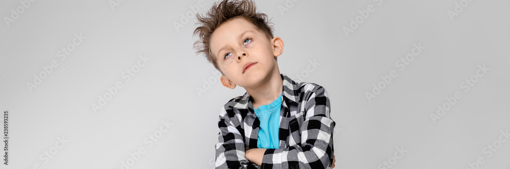 A handsome boy in a plaid shirt, blue shirt and jeans stands on a gray background. The boy folded his arms over his chest. The boy has closed his eyes A handsome boy in a plaid shirt, blue shirt and jeans stands on a gray background. The boy folded his arms over his chest. The boy has closed his eyes