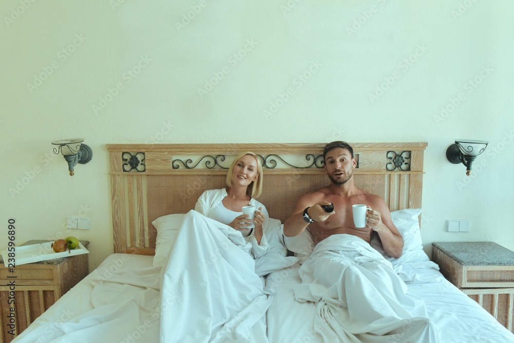 handsome man and beautiful blonde woman lying in bed together having fun watching tv in bedroom, Attractive husband and wife relaxing after sex in the morning, drinking coffee smiling watching comedy
