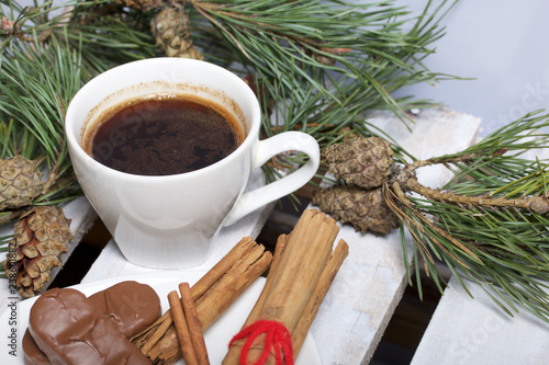 A cup of coffee, Cinnamon sticks and chocolate next to the pine twigs and cones. On a wooden box.