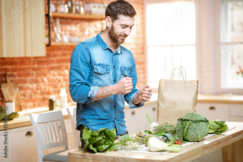 Happy man in blue shirt with excited emotions standing near the table full of green fresh vegetables on the kitchen
