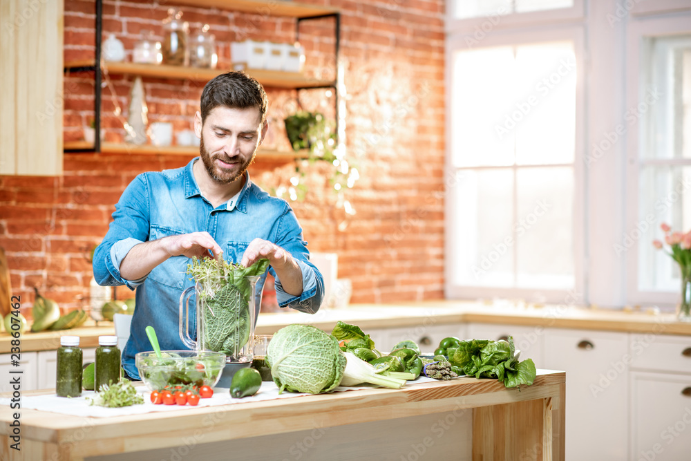 Fototapeta premium Handsome man filling blender with green raw food, making vegetarian smoothie on the kitchen at home