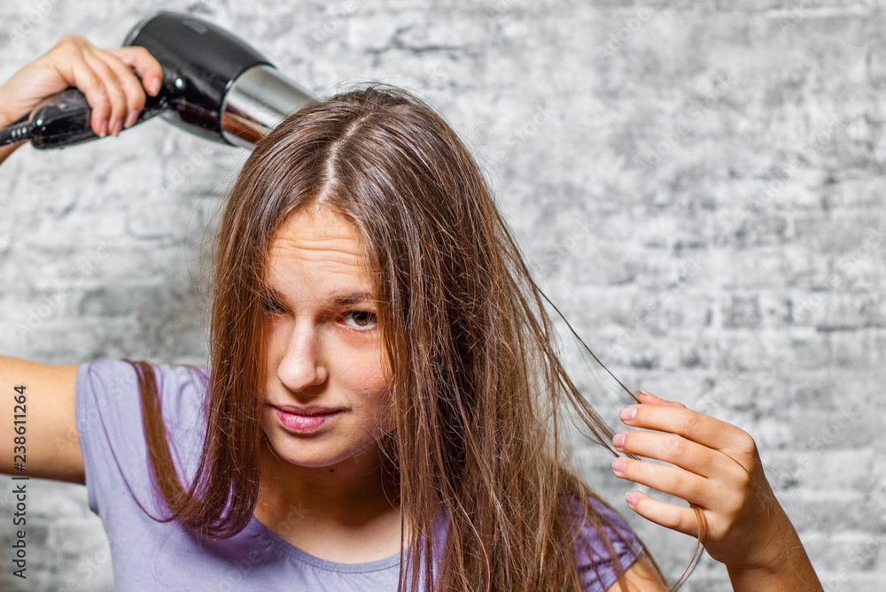 Fototapeta premium portrait of young teenager brunette girl with long hair dries hair with electrical hair dryer on gray wall background