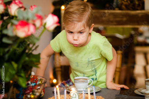 Beautiful adorable six year old boy in green shirt, celebrating his birthday, blowing candles on homemade baked cake, indoor