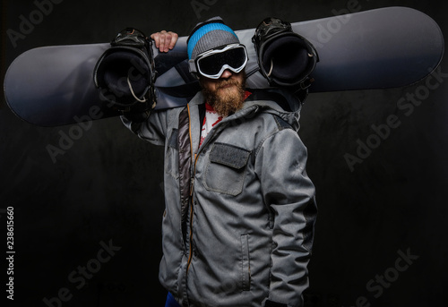 Brutal man with a red beard wearing a full equipment holding a snowboard on his shoulder, isolated on a dark textured background.