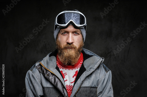 Confident man with a red beard wearing a full equipment for extreme snowboarding, looking at a camera with a serious look, isolated on a dark textured background.