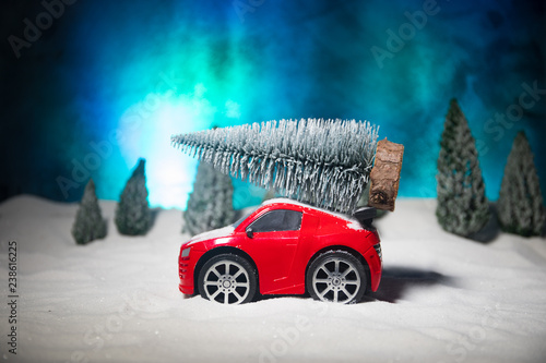 Fototapeta Naklejka Na Ścianę i Meble -  Christmas or New Year concept. Toy car carrying a Christmas tree through the forest in snowfall. Holiday decorated background.