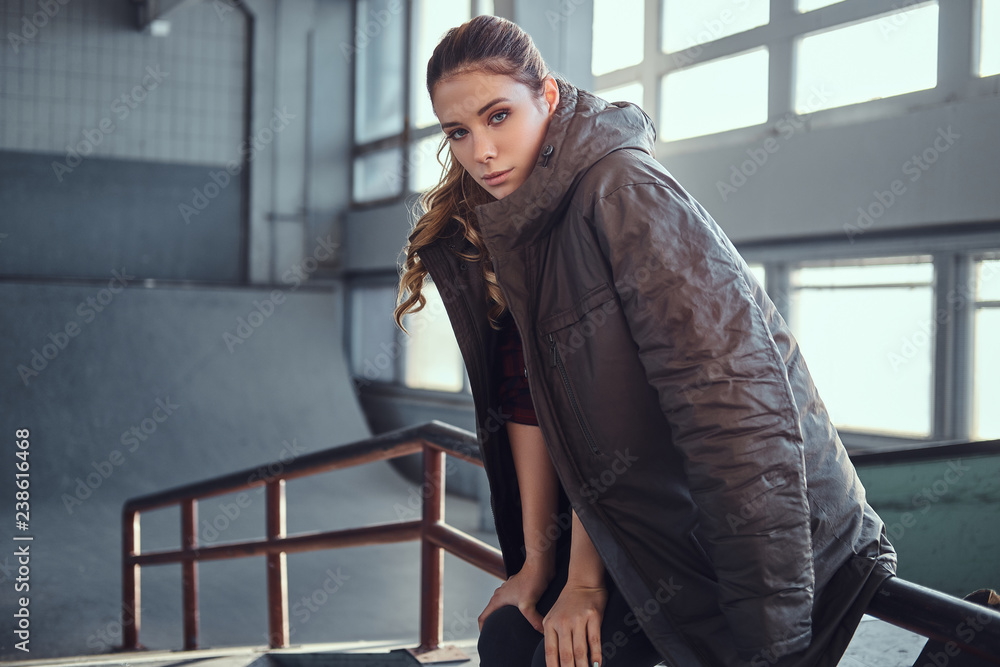 Portrait of a charming young girl with a coat covering his shoulders looking at camera while sitting on a grind rail in skatepark indoors. Portrait of a charming young girl with a coat covering his shoulders looking at camera while sitting on a grind rail in skatepark indoors.