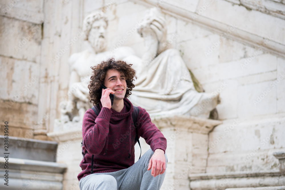 Handsome young man with curly hair in tracksuit talking on his mobile phone in front of Nile God statue in Rome