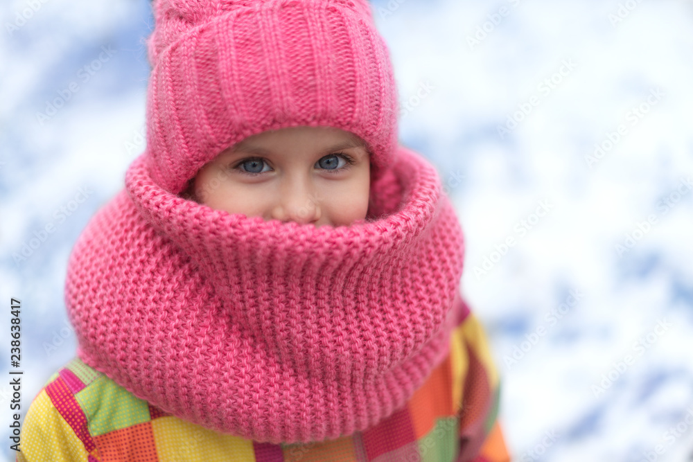 Pretty little girl with smiling eyes is looking through warm hat and snood. Cold winter weather.