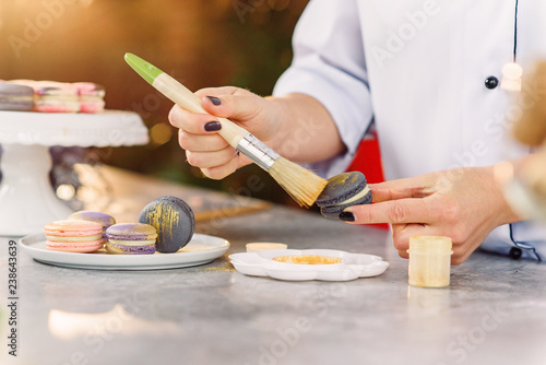 Female confectioner paint with brush a gold food farb on a fresh macarons. Preparing freshly baked macarons.