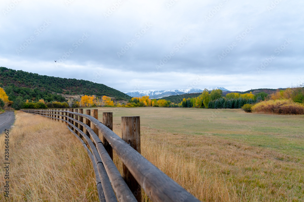 A dirt road lined by a wooden fence curves through fall colored fields