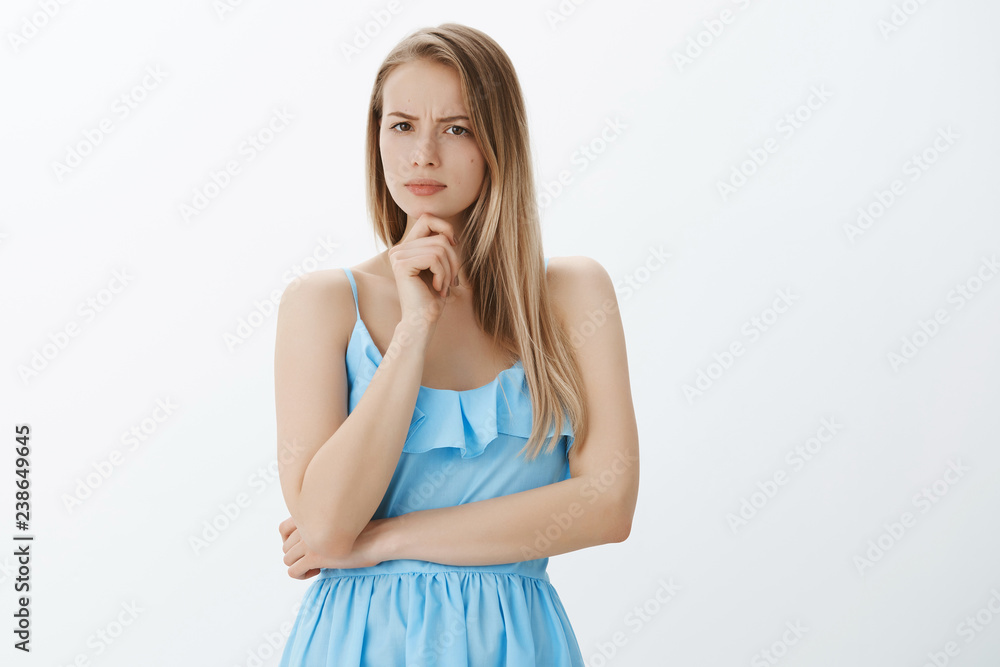 Serious-looking disturbed and suspicious good-looking woman in blue dress with fair hair frowning holding hand on chin, squinting at camera as thinking, being doubtful, expressing disbelief Serious-looking disturbed and suspicious good-looking woman in blue dress with fair hair frowning holding hand on chin, squinting at camera as thinking, being doubtful, expressing disbelief