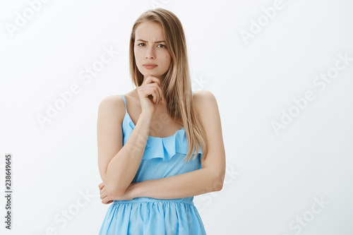 Serious-looking disturbed and suspicious good-looking woman in blue dress with fair hair frowning holding hand on chin, squinting at camera as thinking, being doubtful, expressing disbelief