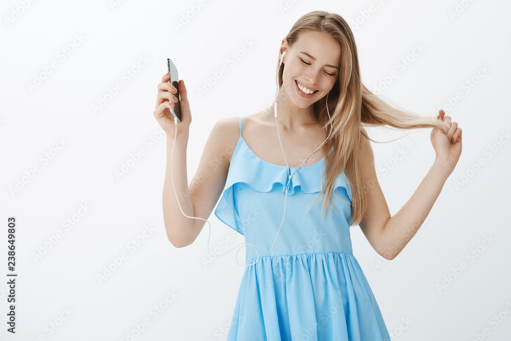 Carefree charismatic young bright european girl with blond hair in blue dress playing with strands looking down with joyful broad smile holding smartphone, listening music in earphones, dancing Carefree charismatic young bright european girl with blond hair in blue dress playing with strands looking down with joyful broad smile holding smartphone, listening music in earphones, dancing