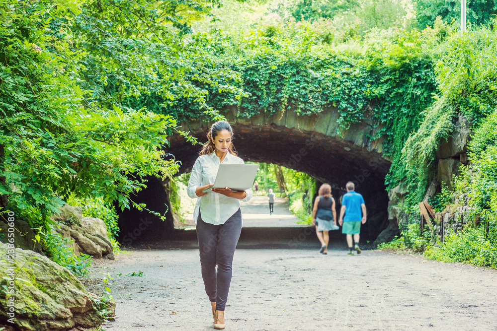 Young East Indian American Woman traveling, working in New York, wearing white shirt, black pants, holding laptop computer, walking at Central Park. Street bridge with green leaves on background..