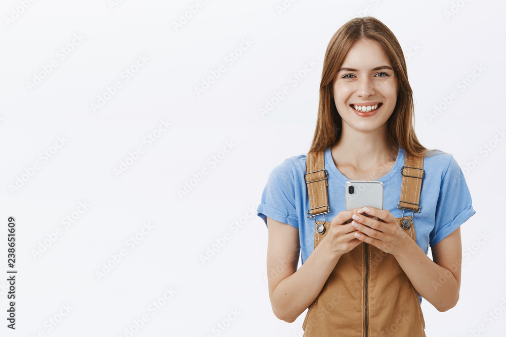 Waist-up shot of charming good-looking young female smartphone user in trendy overalls smiling joyfully with friendly expression holding cellphone edditing photos via new cool app over gray background Waist-up shot of charming good-looking young female smartphone user in trendy overalls smiling joyfully with friendly expression holding cellphone edditing photos via new cool app over gray background