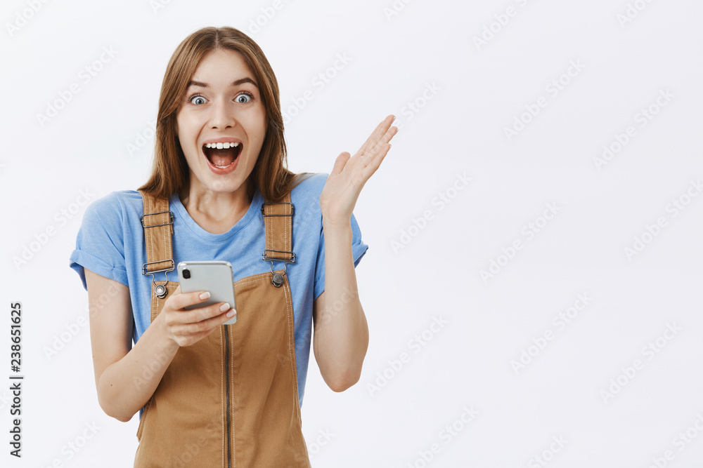 Waist-up shot of excited impressed and surprised charming happy girl in brown overalls raising palm from joy and amazement holding smartphone reacting to awesome astonished news over white wall Waist-up shot of excited impressed and surprised charming happy girl in brown overalls raising palm from joy and amazement holding smartphone reacting to awesome astonished news over white wall