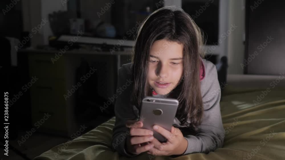 Teenage girl online shopping looks on her smartphone in bed during the night. little teen hood girl writes a message chatting lifestyle in social media messenger night evening indoors smartphone on Teenage girl online shopping looks on her smartphone in bed during the night. little teen hood girl writes a message chatting lifestyle in social media messenger night evening indoors smartphone on