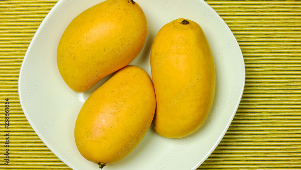Three yellow mangoes on a white plate on a striped background